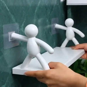Hands installing a white humanoid-shaped wall-mounted floating shelf onto green marble tiles using transparent adhesive pads.
