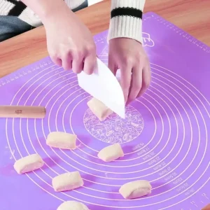 Hands using a dough scraper to cut dough pieces on a bright purple non-stick silicone pastry rolling mat with circular measurement guides.