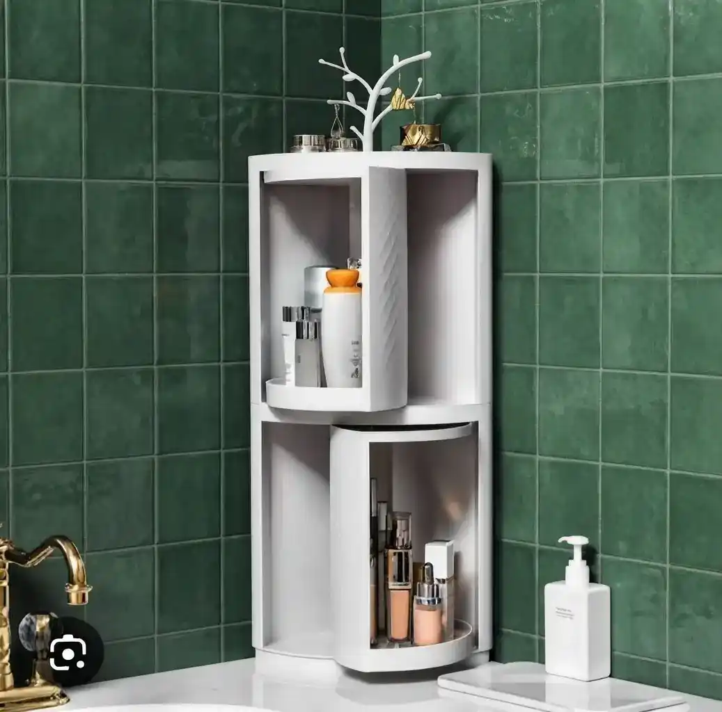 White double-layer 360-degree rotating corner bathroom cabinet with dual storage tiers, cosmetics, and a top jewelry tree holder against green tiles.