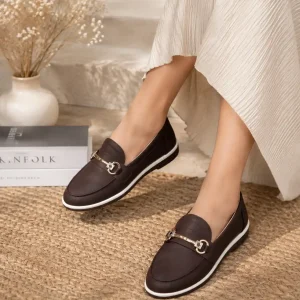 Women's feet wearing dark brown faux leather loafers with gold horsebit buckles styled with a white pleated dress on a woven rug.