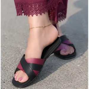 Women's feet wearing maroon and black crisscross flat sandals styled with maroon trousers with lace hem and a gold anklet.