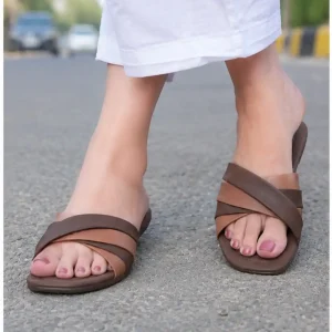 Women's feet wearing multi-tone brown crisscross flat sandals styled with white trousers on an outdoor road.