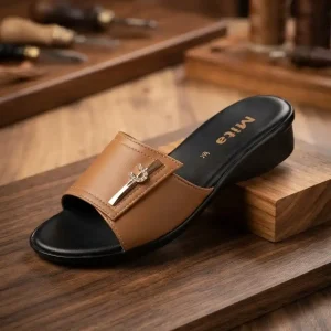 Women's tan low-wedge slip-on slipper with a gold rhinestone buckle displayed on a rustic wooden platform.
